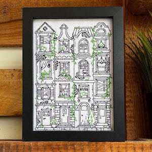 Könnte beinhalten: Ein gerahmtes Kreuzstich-Kunstwerk mit einer detaillierten Darstellung mehrerer Häuser. Die Häuser sind in Schwarz-Weiß mit grünen Akzenten gehalten und in einem schwarzen rechteckigen Rahmen gerahmt. Das Kunstwerk wird auf einem Holzregal ausgestellt.