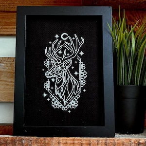 Può includere: Un'opera d'arte in bianco e nero incorniciata con un disegno di testa di cervo. Il disegno bianco è su sfondo nero, in una cornice rettangolare nera. Il cervo ha una luna crescente e stelle. La cornice è su una mensola di legno.