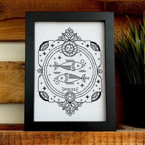 Può includere: Un'opera d'arte a punto croce in bianco e nero incorniciata raffigurante il segno zodiacale dei Pesci. Il disegno include due pesci all'interno di un cerchio, circondati da stelle ed elementi decorativi. La parola "PISCES" è ricamata sotto.