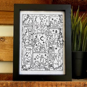 Op de afbeelding: Zwartgelijste kruissteekkunst met diverse drankflessen, kaarsen en hemelse elementen. De woorden "MAGIC", "LOVE" en "POISONS" zijn zichtbaar. Het kunstwerk wordt tentoongesteld op een houten plank met een potplant.
