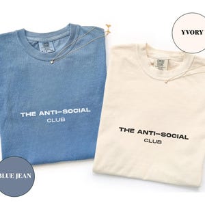 K&ouml;nnte beinhalten: Zwei T-Shirts, eines in Blue Jean und eines in Ivory, werden gezeigt. Auf jedem Shirt steht in Wei&szlig; "THE ANTI-SOCIAL CLUB". Auf jedem Shirt liegt eine zarte goldene Halskette mit einem kleinen Anh&auml;nger. Der Markenname "Sisters Project & Co." steht unten.