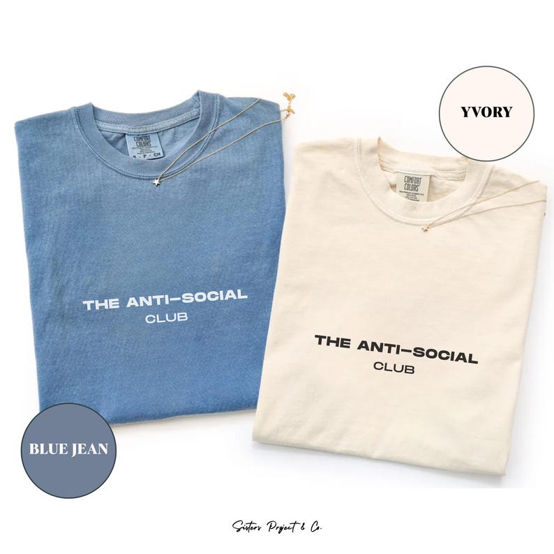 K&ouml;nnte beinhalten: Zwei T-Shirts, eines in Blue Jean und eines in Ivory, werden gezeigt. Auf jedem Shirt steht in Wei&szlig; "THE ANTI-SOCIAL CLUB". Auf jedem Shirt liegt eine zarte goldene Halskette mit einem kleinen Anh&auml;nger. Der Markenname "Sisters Project & Co." steht unten.
