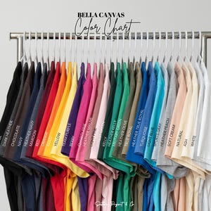 K&ouml;nnte beinhalten: Eine Farbkarte zeigt eine Vielzahl von Bella Canvas T-Shirts in verschiedenen Farben, darunter Schwarz, Marineblau, Rot, Gelb und Wei&szlig;. Die Shirts h&auml;ngen an einem Metallgestell, wobei jede Farbe zur einfachen Referenz gekennzeichnet ist. Der Text "BELLA CANVAS Color Chart" steht oben.
