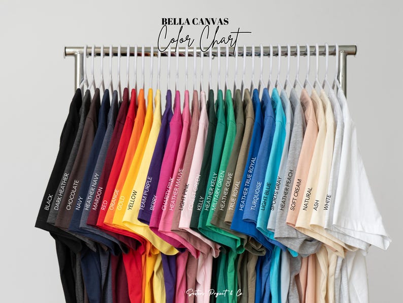 K&ouml;nnte beinhalten: Eine Farbkarte zeigt eine Vielzahl von Bella Canvas T-Shirts in verschiedenen Farben, darunter Schwarz, Marineblau, Rot, Gelb und Wei&szlig;. Die Shirts h&auml;ngen an einem Metallgestell, wobei jede Farbe zur einfachen Referenz gekennzeichnet ist. Der Text "BELLA CANVAS Color Chart" steht oben.