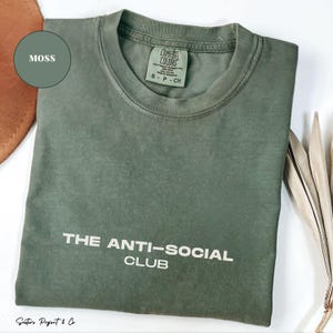 K&ouml;nnte beinhalten: Ein moosgr&uuml;nes T-Shirt mit dem wei&szlig;en Text "THE ANTI-SOCIAL CLUB". Das Shirt hat einen Rundhalsausschnitt und ein Etikett mit der Aufschrift "COMFORT COLORS". Das Shirt ist gefaltet. Ein kleines Logo befindet sich in der Ecke.