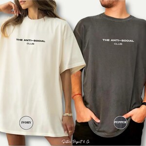 K&ouml;nnte beinhalten: Zwei &uuml;bergro&szlig;e T-Shirts, eines in Elfenbein und eines in Pfeffer, jeweils mit dem Text "THE ANTI-SOCIAL CLUB" in Schwarz bedruckt. Das elfenbeinfarbene Shirt ist links und das pfefferfarbene Shirt rechts. Der Markenname "Sisters Project & Co." steht unten.