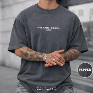 K&ouml;nnte beinhalten: Pfefferfarbenes, &uuml;bergro&szlig;es T-Shirt mit dem wei&szlig;en Text "THE ANTI-SOCIAL CLUB". Das Shirt hat einen Vintage-Look. Das Model tr&auml;gt ein silbernes Armband und einen Ring.