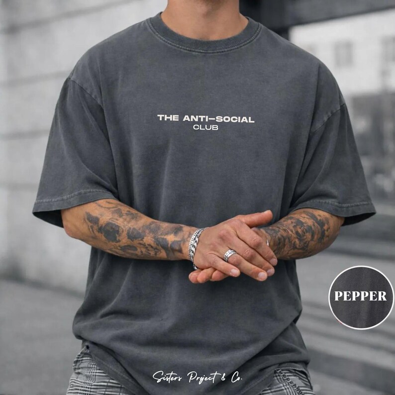 K&ouml;nnte beinhalten: Pfefferfarbenes, &uuml;bergro&szlig;es T-Shirt mit dem wei&szlig;en Text "THE ANTI-SOCIAL CLUB". Das Shirt hat einen Vintage-Look. Das Model tr&auml;gt ein silbernes Armband und einen Ring.