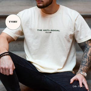K&ouml;nnte beinhalten: Cremefarbenes T-Shirt mit dem Text "THE ANTI-SOCIAL CLUB" in Dunkelgr&uuml;n. Das kurz&auml;rmelige Shirt ist aus einem weichen Material. Das Model tr&auml;gt eine schwarze Jeans und ein schwarzes Armband.
