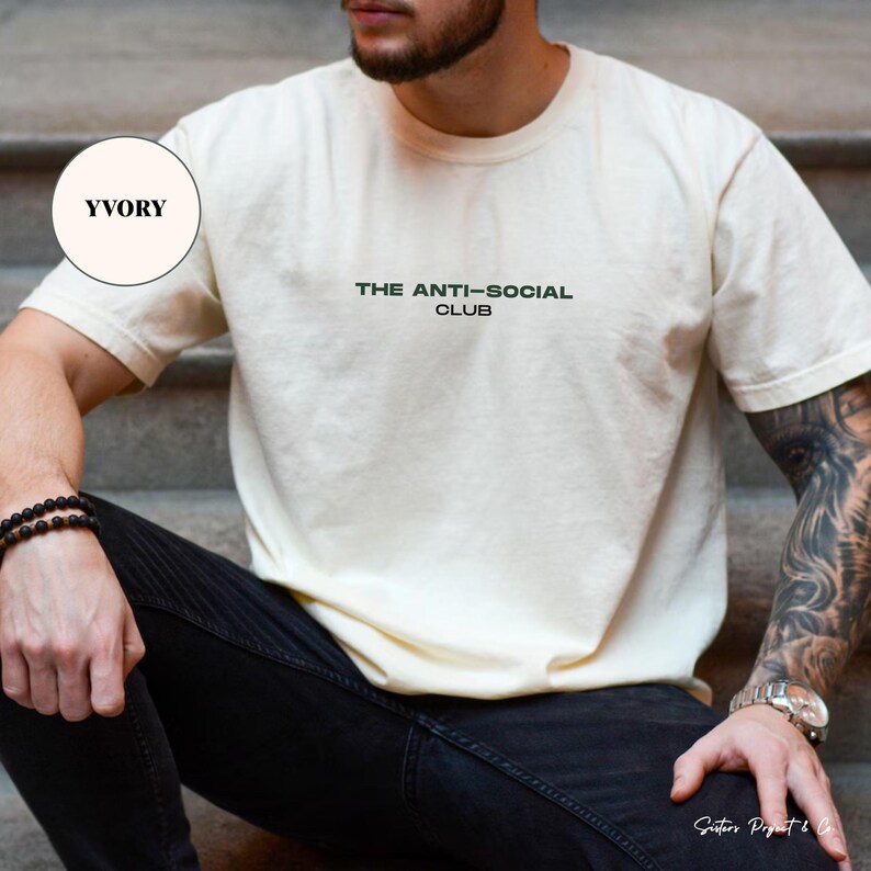 K&ouml;nnte beinhalten: Cremefarbenes T-Shirt mit dem Text "THE ANTI-SOCIAL CLUB" in Dunkelgr&uuml;n. Das kurz&auml;rmelige Shirt ist aus einem weichen Material. Das Model tr&auml;gt eine schwarze Jeans und ein schwarzes Armband.