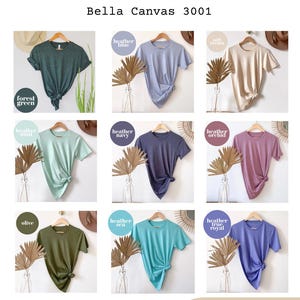 K&ouml;nnte beinhalten: Farbkarte mit Bella Canvas 3001 T-Shirts in verschiedenen Farben, darunter Waldgr&uuml;n, Heather Blue und Olive. Jedes Shirt wird auf einem Kleiderb&uuml;gel mit Knotendetail pr&auml;sentiert. Der Text "More colors available" ist ebenfalls vorhanden.