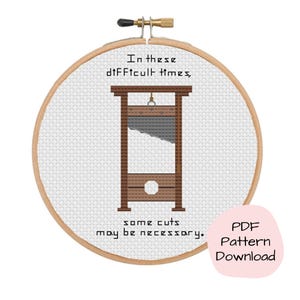 May include: Cross-stitch embroidery in a wooden hoop features a brown guillotine design with the text "In these difficult times, some cuts may be necessary." A pink speech bubble says "PDF Pattern Download."