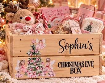 May include: A wooden crate labeled Sophia's Christmas Eve Box, decorated with fairy and tree illustrations. Inside are a plush reindeer, a book titled Santa's Little Helper, a pink tumbler, a bag of hot chocolate, and fuzzy socks.