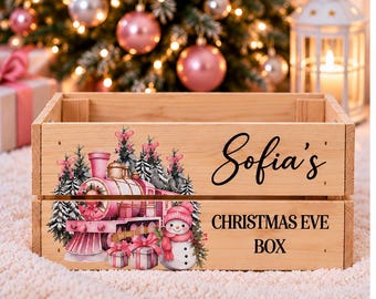 May include: A light-colored wooden crate featuring a printed graphic of a pink train, a snowman, and wrapped gifts. The text "Sofia's" appears in black script above the printed text "CHRISTMAS EVE BOX" in a bold, black sans-serif font.