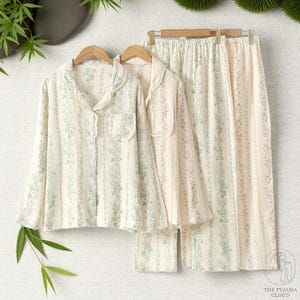 May include: Cream-colored pajama set with a floral stripe pattern in green and pink. The set includes a long-sleeved button-down shirt with a notched collar and a pair of matching pants. The pajamas are displayed on wooden hangers.