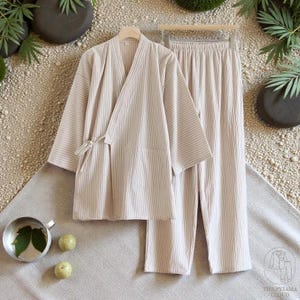 Peut inclure: Un ensemble de pyjama beige à rayures, comprenant un haut de style enveloppant avec une fermeture à nouer et un pantalon assorti. L'ensemble est présenté sur un cintre, mettant en valeur les tons doux et neutres et le motif à rayures verticales. Le pyjama est en tissu léger.