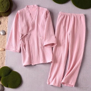 May include: A light pink pajama set, comprising a long-sleeved wrap top with a tie closure and matching trousers. The fabric has a textured, crinkled appearance. The set is displayed on a neutral surface.