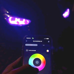 Può includere: Uno schermo di smartphone mostra un'interfaccia a ruota cromatica per controllare l'illuminazione RGB. Il telefono è tenuto in mano, con i fari di un'auto illuminati di viola sullo sfondo. Il testo "ELK-BLEDWM DF" è visibile.