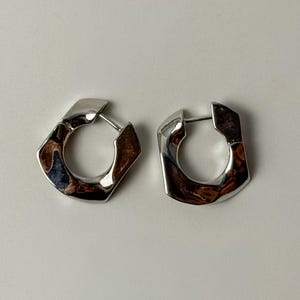 May include: A pair of silver-toned hoop earrings with a distinct, irregular, and angular geometric shape. The metal surface is highly polished and reflective, creating a smooth, fluid appearance with visible highlights and dark shadows.