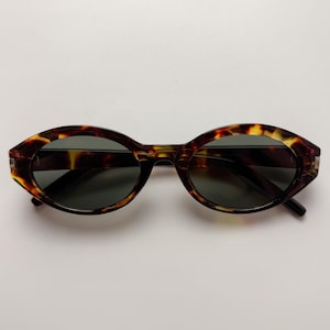 May include: Oval sunglasses with a tortoiseshell design. The frames are brown and yellow, and the lenses are dark green. The sunglasses are set against a white background.