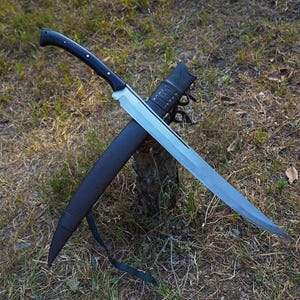 May include: A large, silver-bladed sword with a black handle and a black sheath. The sword is crossed over the sheath, resting on a bed of grass and leaves. The handle has a curved design with small white accents.