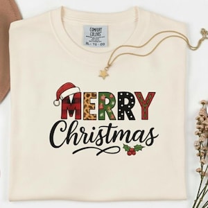 Puede incluir: Sudadera de cuello redondo color crema con las palabras "MERRY Christmas" en un diseño festivo. La palabra "MERRY" presenta un gorro de Papá Noel y un estampado de leopardo. Un collar dorado con un colgante de estrella está sobre la camisa.