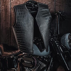 Könnte beinhalten: Eine schwarze ärmellose Weste mit einer strukturierten Krokodilleder-Optik. Die Weste hat einen V-Ausschnitt und eine strukturierte Form und liegt auf dem Kraftstofftank eines schwarzen Motorrads mit sichtbaren Motorteilen und Lenker.
