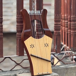 Może przedstawiać: Drewniana lira z ciemnobrązową ramą i jasnobrązową płytą rezonansową. Płyta rezonansowa posiada dwa wycięcia w kształcie gwiazd i ciemnobrązowy drążek. Instrument posiada wiele strun i spoczywa na kamiennej powierzchni.