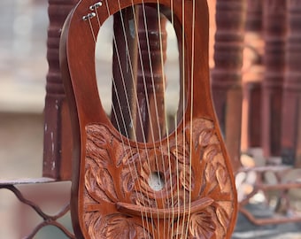 May include: A wooden lyre with a carved floral design. The instrument has a dark brown finish, with strings stretched across the sound hole. The lyre is resting on a concrete surface, with a blurred background of a fence.