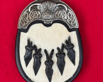 Op de afbeelding: Een traditionele Schotse sporran met een zilverkleurige metalen bovenkant met een Highland koeienontwerp en Keltisch knoopwerk. De sporran heeft een witte vacht body, zwarte leren afwerking en vijf zwarte leren kwasten.