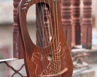 Puede incluir: Una lira de madera con un diseño tallado de una figura tocando un arpa. El instrumento tiene 10 cuerdas y está hecho de madera marrón oscuro. La lira descansa sobre una superficie de piedra.