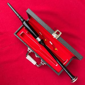 Puede incluir: Un puntero de gaita negro con detalles plateados, presentado en un estuche negro abierto con interior de terciopelo rojo. El estuche está sobre un fondo rojo, mostrando el instrumento.