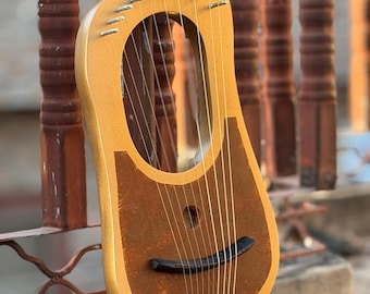 Puede incluir: Una lira de madera con marco dorado y caja de resonancia marrón. El instrumento tiene cuerdas tensadas sobre el cuerpo y un puente curvo. La lira descansa sobre una superficie de piedra.