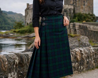 May include: A woman wearing a black button-down shirt and a green and navy plaid kilt. The kilt has two buckle closures. She is wearing black boots. A stone bridge and a castle are in the background.