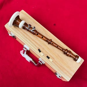 Könnte beinhalten: Eine Holzoboe mit weißem Schallbecher und Akzenten, die auf einem hellen Holzkoffer mit silberner Hardware liegt. Zwei Blätter sind neben der Oboe platziert. Der Hintergrund ist einfarbig rot.