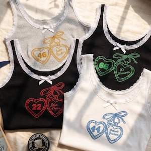 May include: Four ribbed tank tops with white lace trim and a small centre bow. Each features an embroidered design of two hearts tied with a bow, containing a number and a name: 46 John Tucker, 66 D. Laurentis, 22 John Logan, and 44 Garrett Graham.