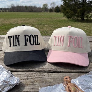 May include: Two baseball caps with off-white crowns and the text TIN FOIL embroidered on the front. One cap features a black brim and black lettering, while the other cap features a light pink brim and light pink lettering, both with a button top.