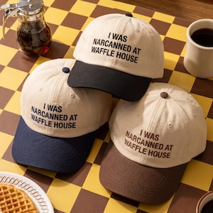 May include: Three beige baseball caps with the text "I WAS NARCANNED AT WAFFLE HOUSE" in black. Each cap has a different coloured brim: black, navy blue, and brown. The caps are arranged on a checkered yellow and brown surface.