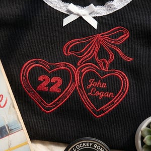 May include: A black ribbed shirt featuring a white lace collar with a small white bow. The shirt has red embroidery of two heart-shaped lockets tied with a red ribbon. One heart contains the number 22, and the other contains the text John Logan.