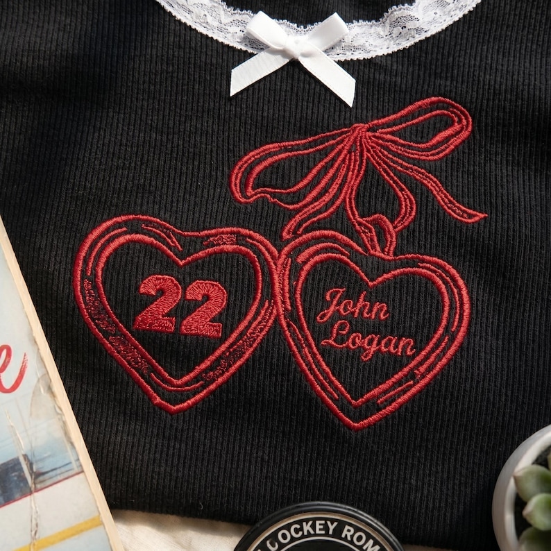 May include: A black ribbed shirt featuring a white lace collar with a small white bow. The shirt has red embroidery of two heart-shaped lockets tied with a red ribbon. One heart contains the number 22, and the other contains the text John Logan.
