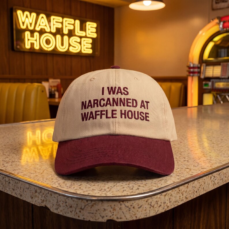 May include: A beige baseball cap with a maroon peak. The cap displays the maroon text "I WAS NARCANNED AT WAFFLE HOUSE". The background shows a Waffle House sign and a diner interior.