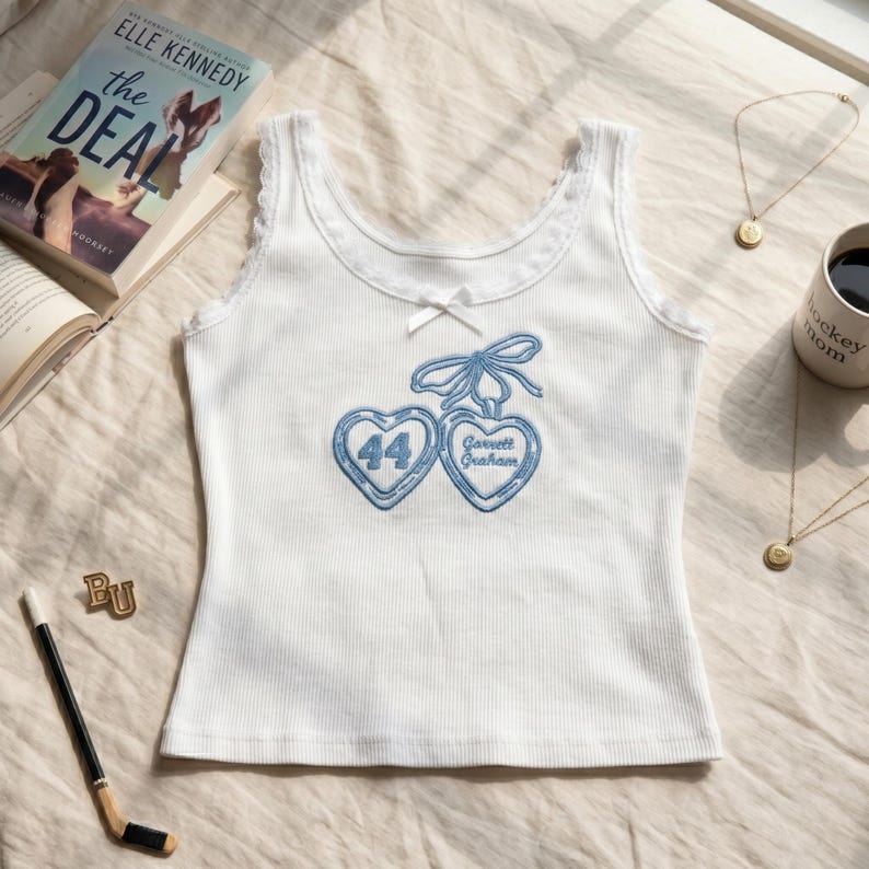 May include: A white ribbed vest top featuring a small white bow at the centre neckline and lace trim along the straps. The front displays blue embroidery of two heart-shaped pendants connected by a bow, with the number 44 and the text Garrett Graham.
