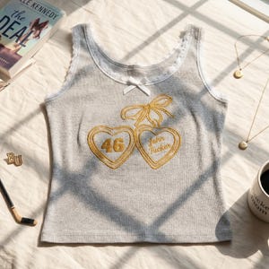 May include: A grey ribbed vest top featuring white lace trim along the neckline and armholes, a small white bow at the centre, and an embroidered design of two gold hearts connected by a bow, with the number 46 and the text John Tucker inside.