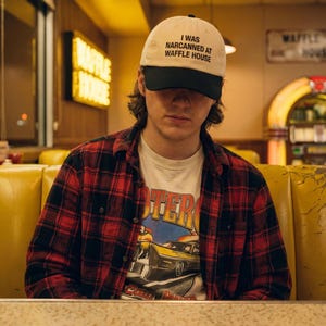 May include: A person wearing a cream baseball cap with the text "I WAS NARCANNED AT WAFFLE HOUSE" in black. They are also wearing a red and black plaid shirt over a graphic tee. The background shows a diner setting.