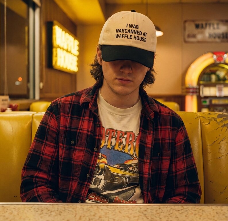 May include: A person wearing a cream baseball cap with the text "I WAS NARCANNED AT WAFFLE HOUSE" in black. They are also wearing a red and black plaid shirt over a graphic tee. The background shows a diner setting.