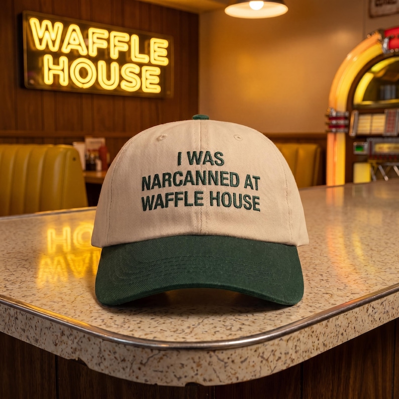May include: A beige and green baseball cap with the text "I WAS NARCANNED AT WAFFLE HOUSE" embroidered in green. The cap has a curved brim and is on a countertop. A neon sign that says "WAFFLE HOUSE" is in the background.