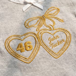 May include: A light grey ribbed fabric garment featuring an embroidered gold design of two hearts connected by a bow. The left heart contains the number 46, and the right heart contains the embroidered text John Tucker. A white ribbon is at the top.