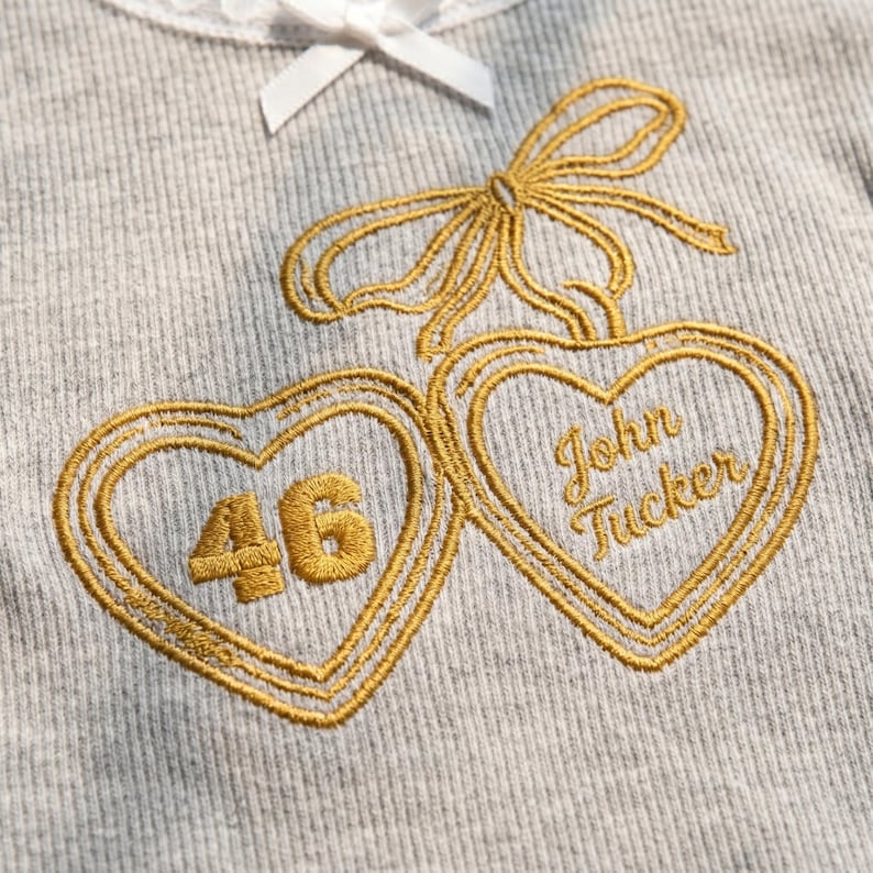 May include: A light grey ribbed fabric garment featuring an embroidered gold design of two hearts connected by a bow. The left heart contains the number 46, and the right heart contains the embroidered text John Tucker. A white ribbon is at the top.