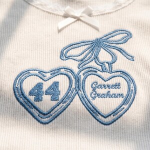 May include: A white ribbed shirt featuring a small white bow at the neckline and blue embroidery of two heart-shaped cherries. The left heart contains the number 44, and the right heart contains the text Garrett Graham in blue stitching.