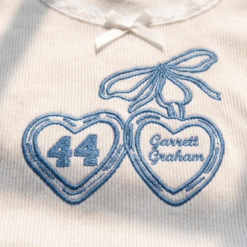 May include: A white ribbed shirt featuring a small white bow at the neckline and blue embroidery of two heart-shaped cherries. The left heart contains the number 44, and the right heart contains the text Garrett Graham in blue stitching.